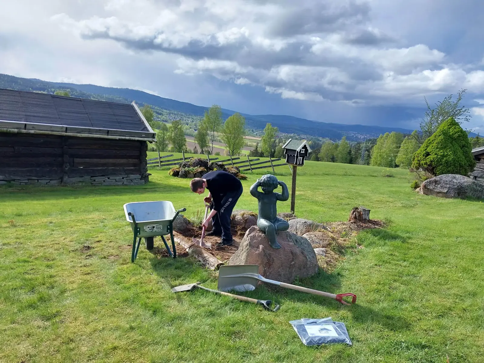 En person graver i jorda ved en trillebår, med grønne åser bak.