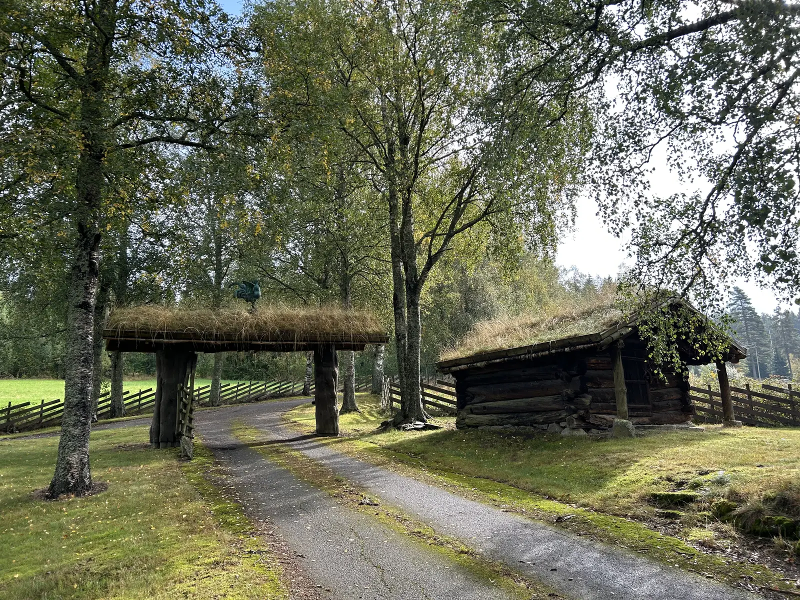 En grusvei som går under en tømmerport med torvtak, med trær og en tømmerhytte langs veien.