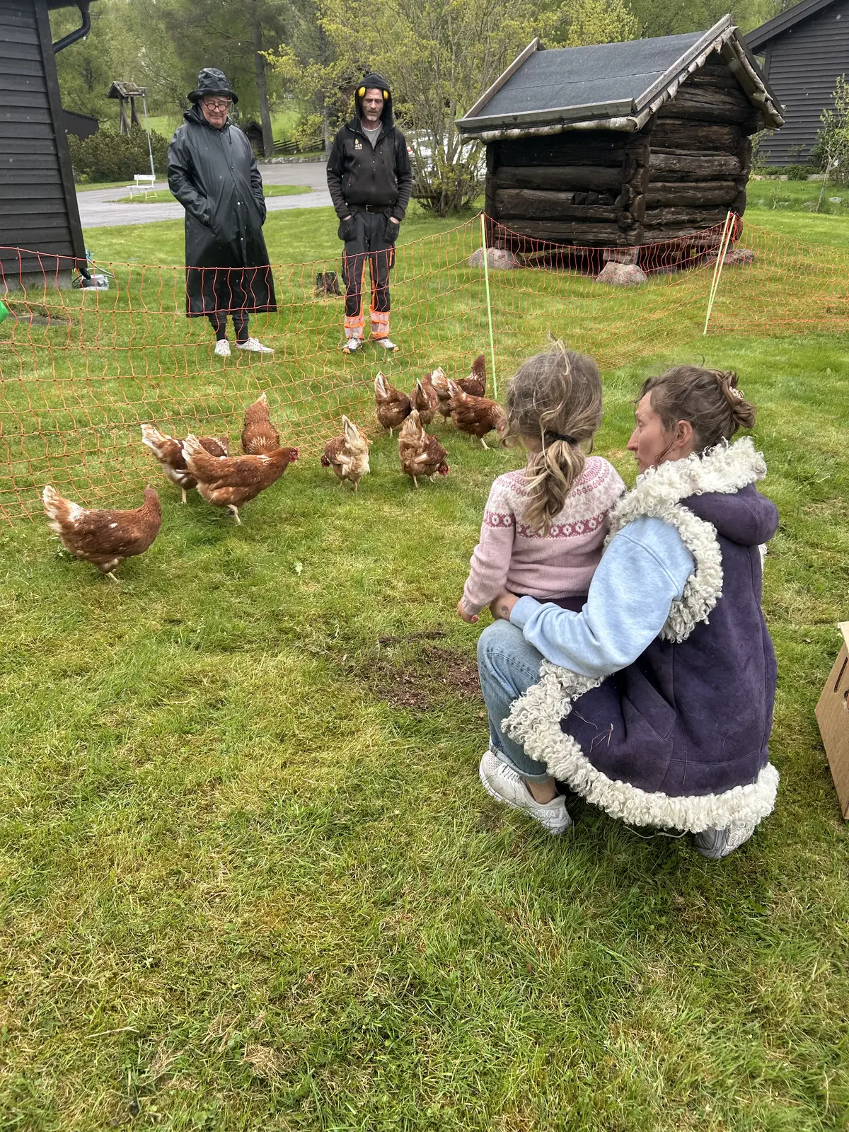 En kvinne sitter på hælene med et barn og ser på en flokk høner bak et oransje nett, med to menn og et gammelt tømmerskjul bak.