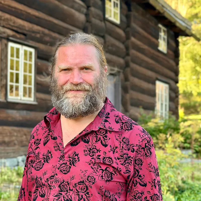 Portrett av Tharald Nustad i rød mønstret skjorte foran det gamle tømmerhuset.