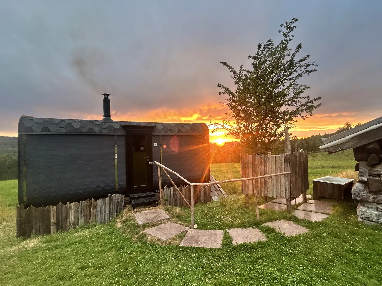 A black barrel sauna with smoke rising from its chimney against an orange evening sky.