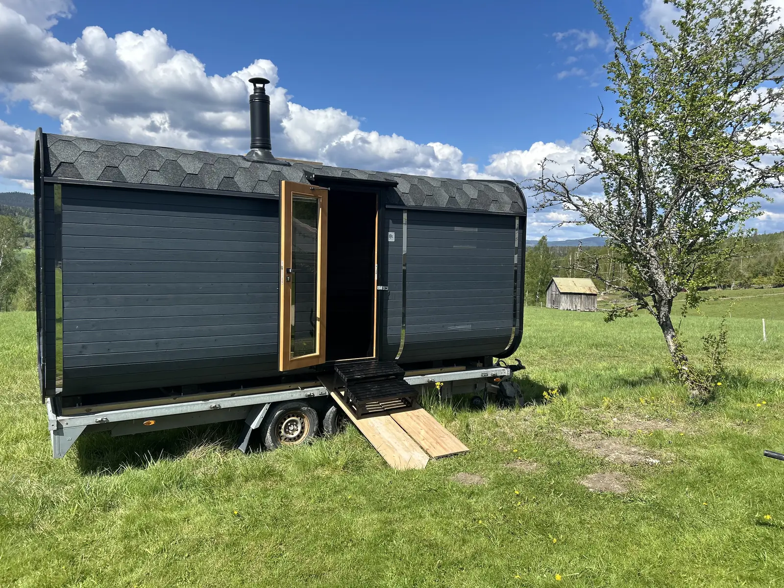 Den svarte tønnebadstua står på en henger i en grønn eng under blå himmel.