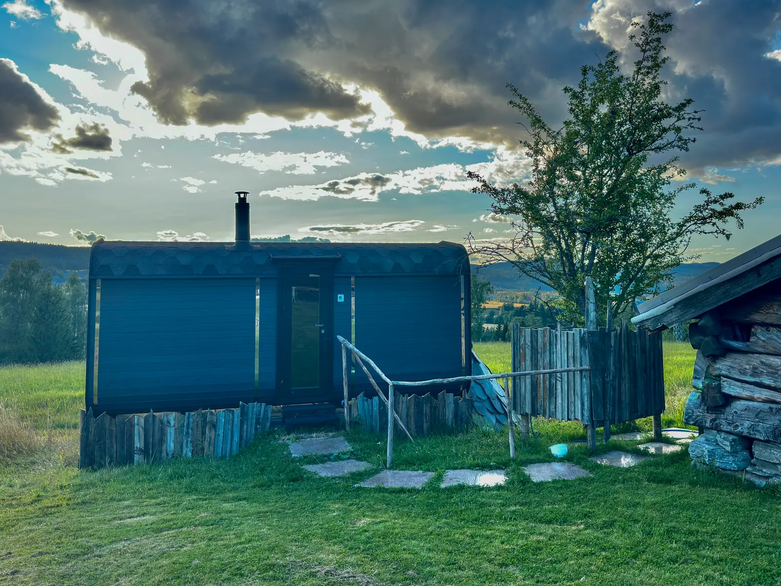 The black sauna with its chimney and wooden steps in a field under a dramatic evening sky.