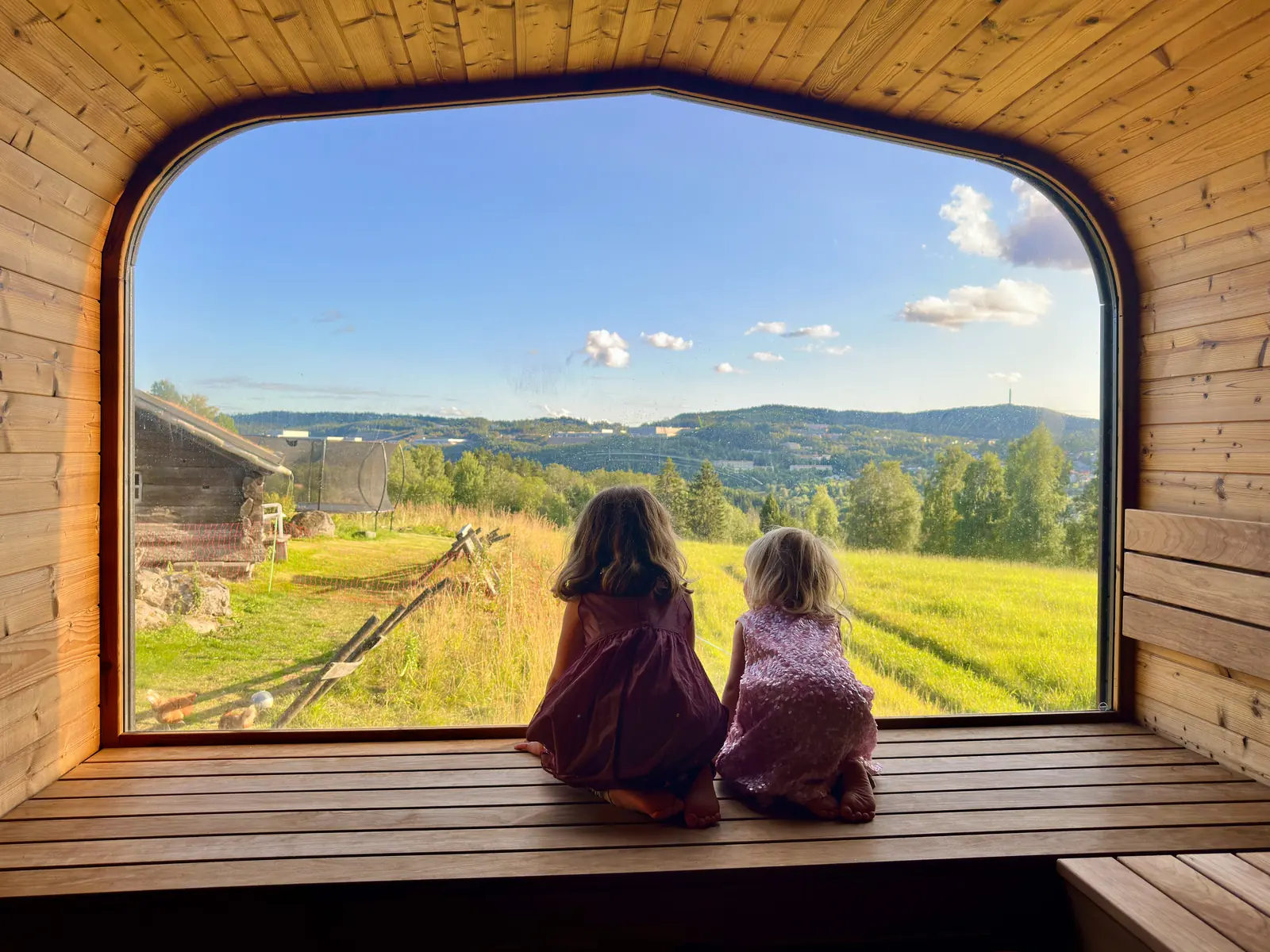 Two children sitting on the sauna bench, looking out through the large window over fields and the valley.