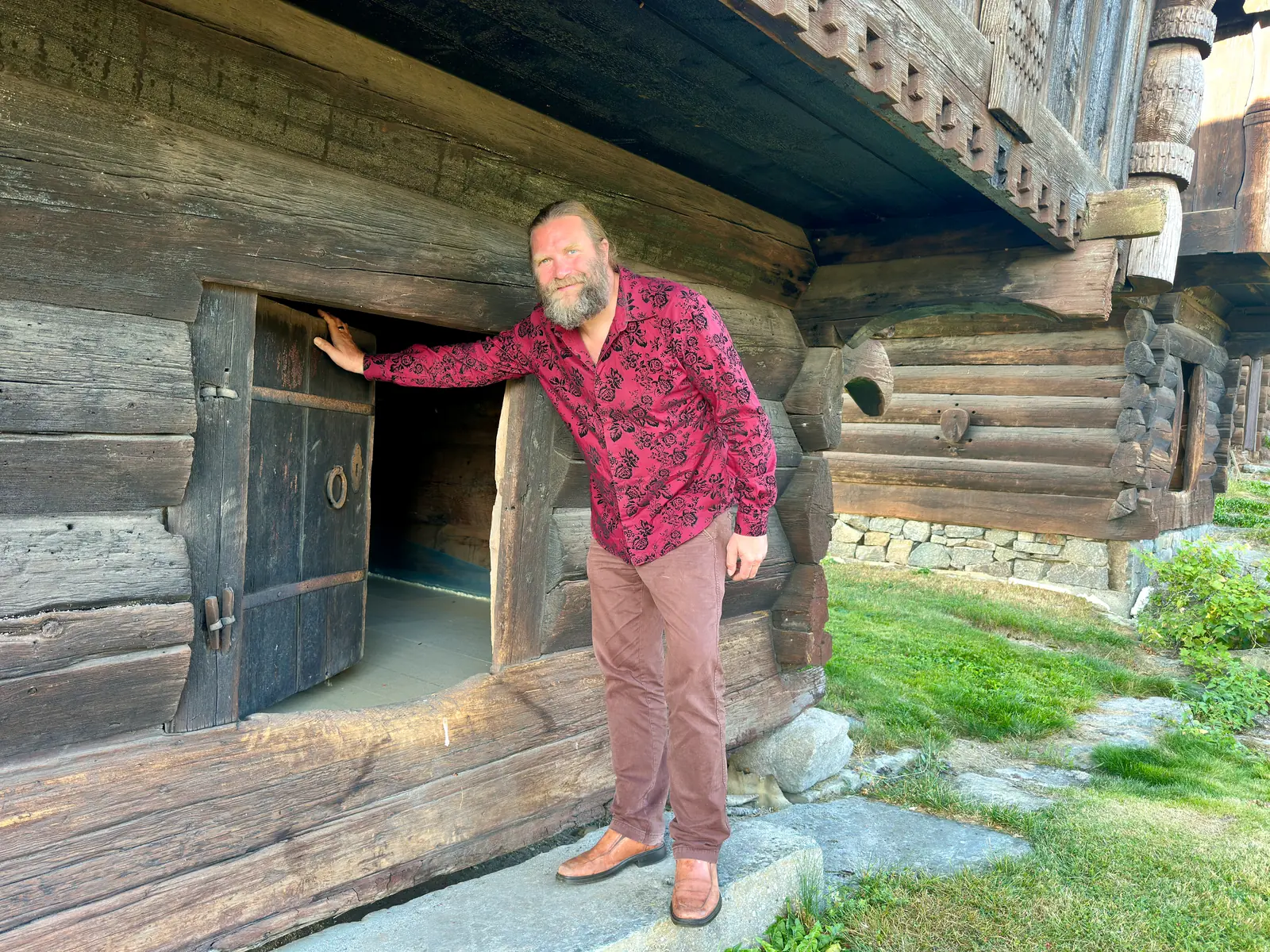 A bearded man in a red shirt standing at the low door of an old log storehouse with a carved loft above.