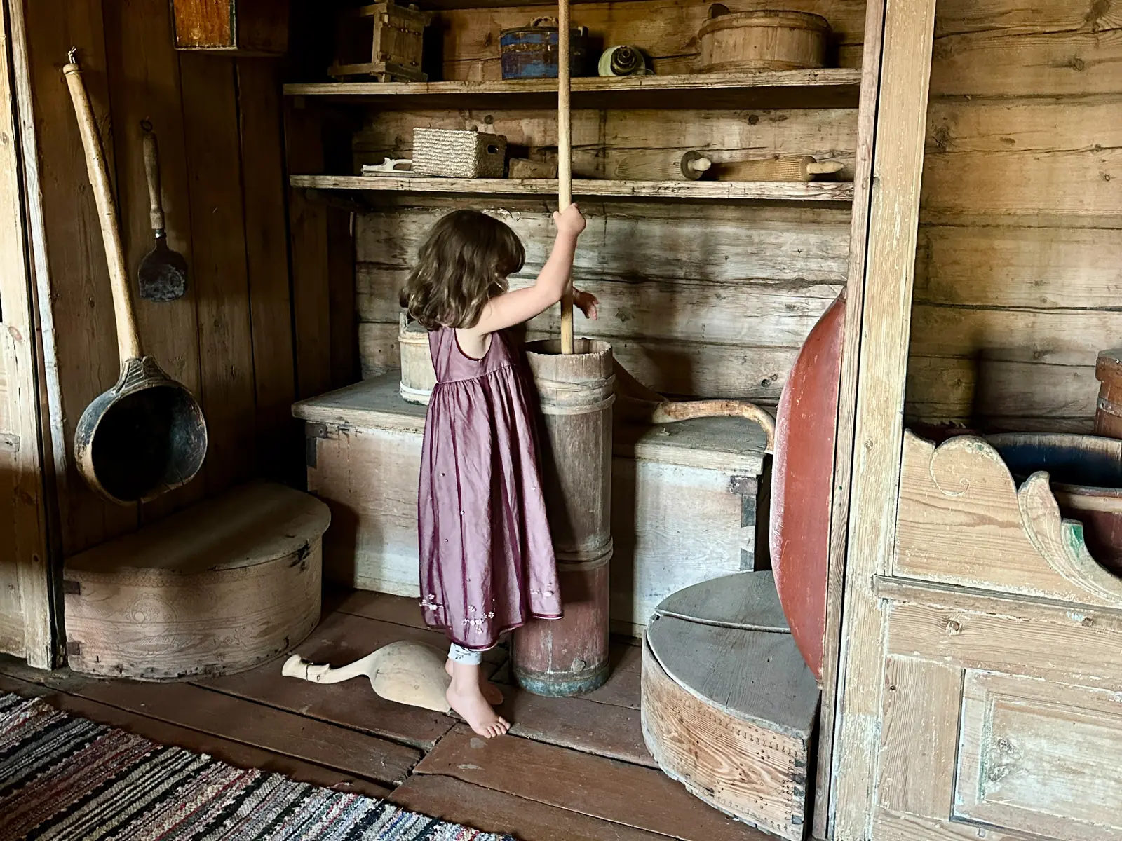 Et barn i lilla kjole kinner smør i en gammel trekinne ved hyller med treredskaper.