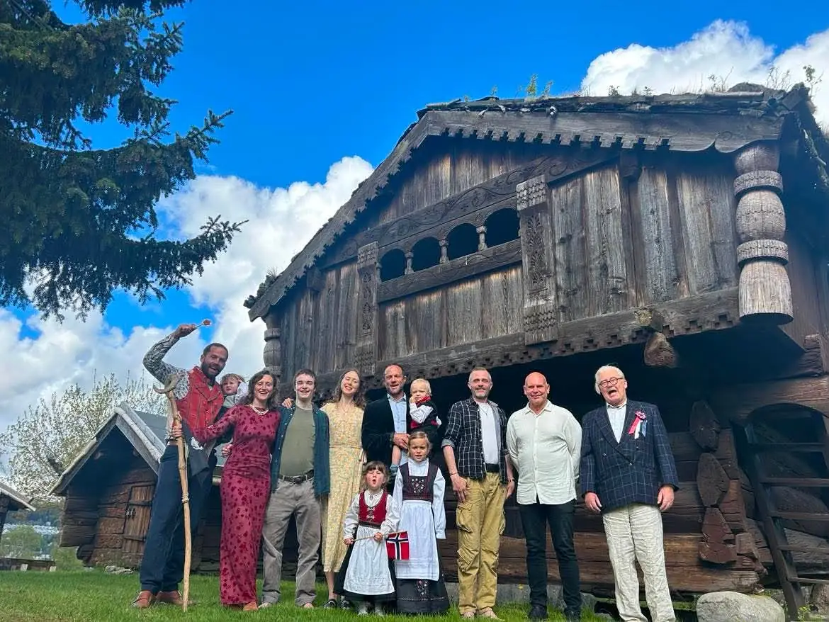 En stor gruppe voksne og barn, noen i bunad, står foran en gammel tømmerbygning.