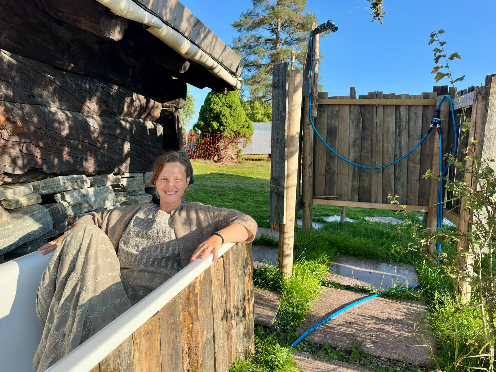 A woman smiling from a wooden cold water tub beside an old log building.