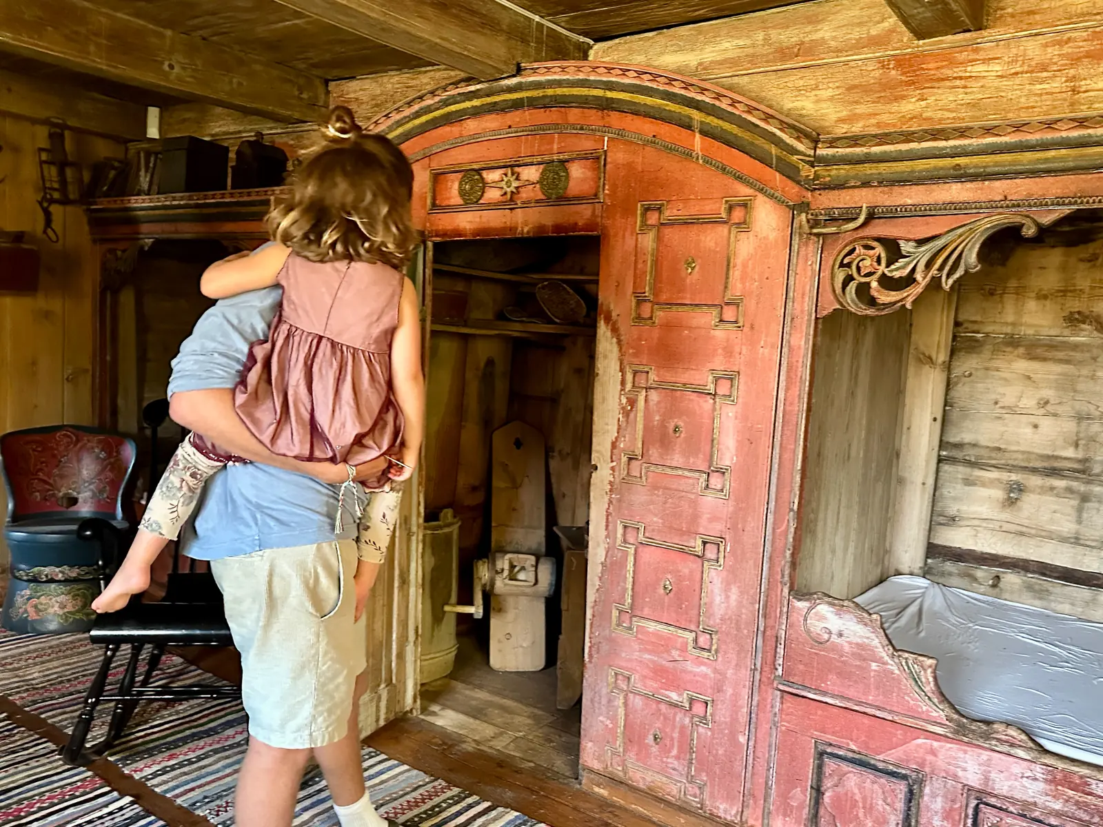 A person carrying a child past a red painted cupboard and built in bed in an old timber room.