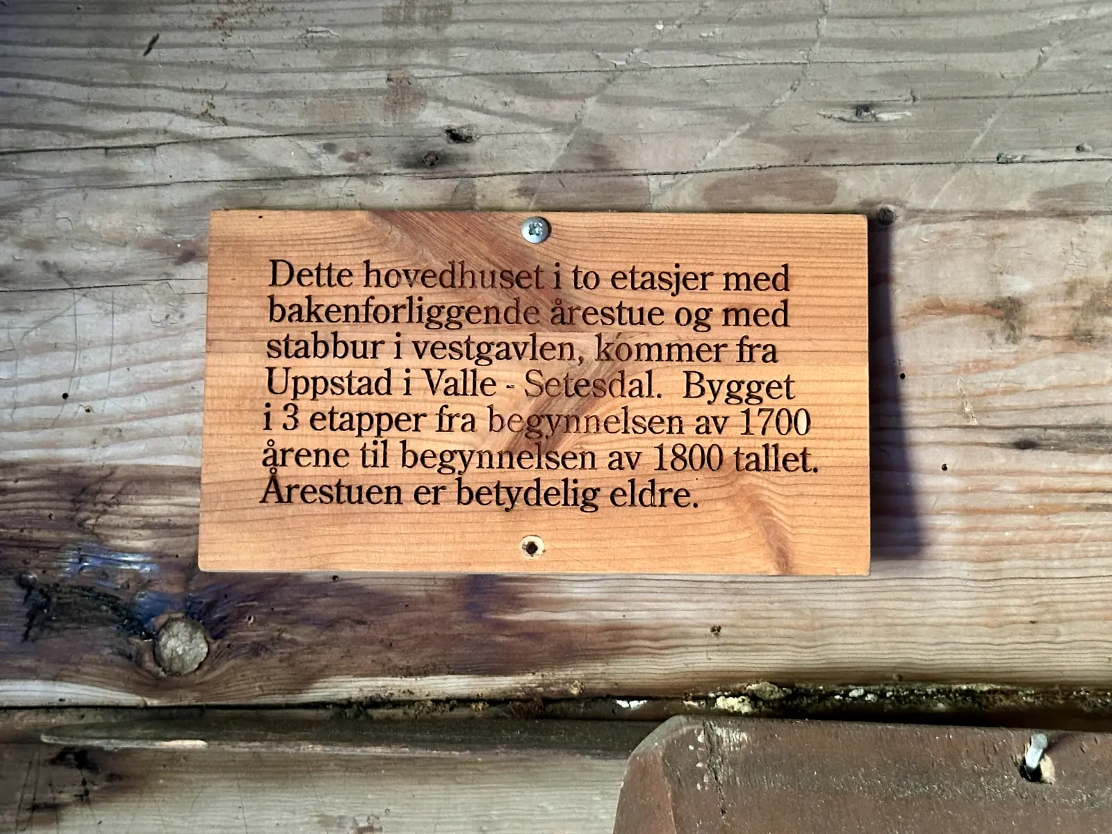 A carved wooden sign on a log wall explaining that the main house comes from Uppstad in Valle, Setesdal, built in three stages from the early 1700s to the early 1800s.